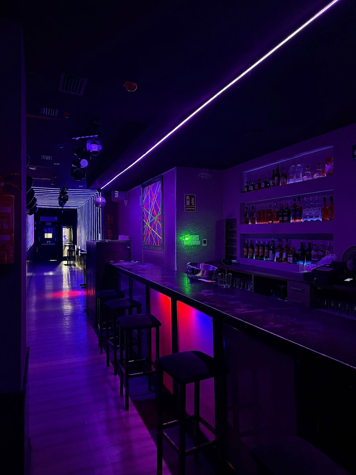 Sala Purple en Barcelona, barra larga con neones morados, estanterías de botellas iluminadas, taburetes altos y mural fluorescente